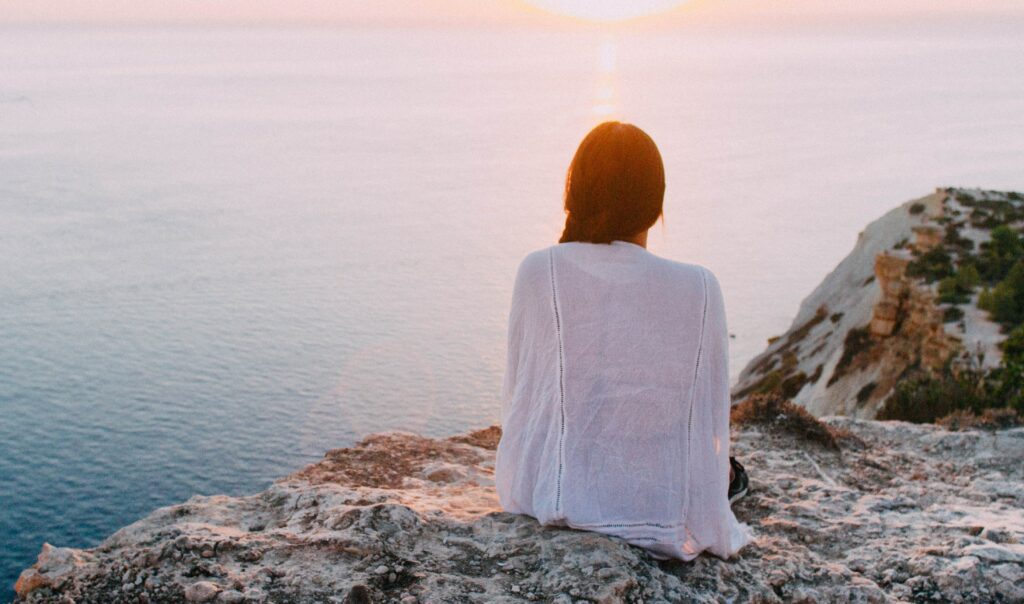 A woman is sitting on a rocky beach looking at the water to represent meth addiction and meth addiction rehab. 